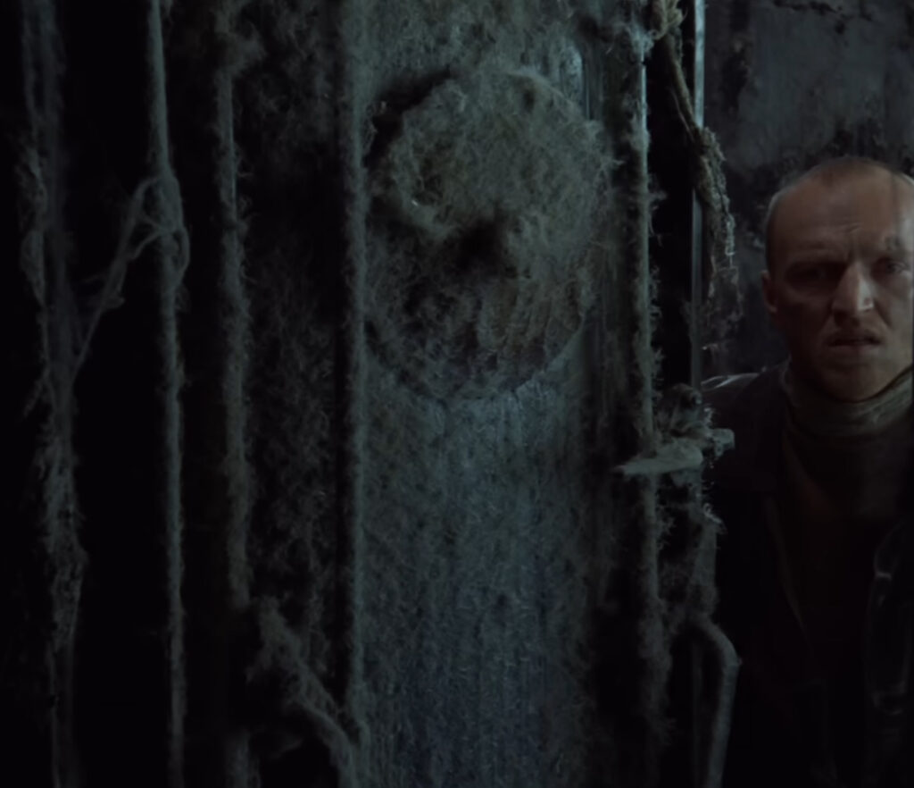 Still from Andrei Tarkovsky's Stalker: the shaven-headed Stalker peers past a moss and cobweb covered pillar in the damp, overgrown Zone.